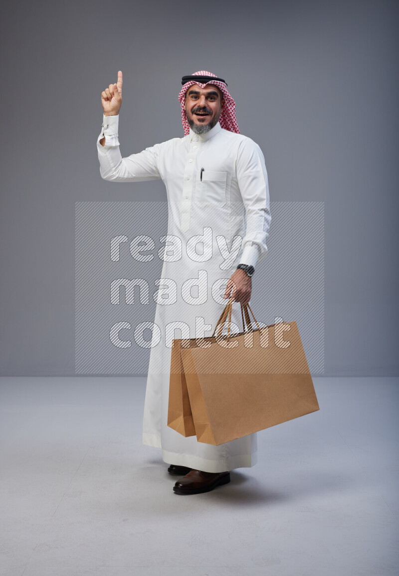 Saudi man Wearing Thob and red Shomag standing holding shopping bag on Gray background