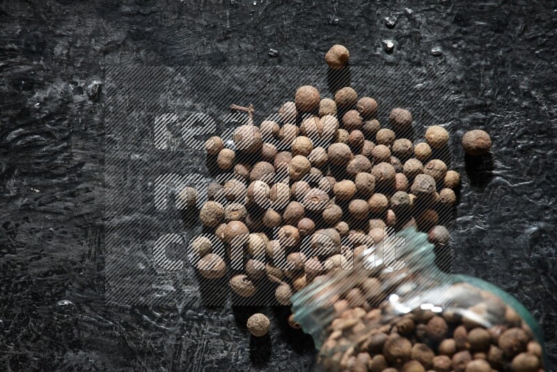 A flipped glass spice jar full of allspice whole balls and the balls spilled out of it on a textured black flooring