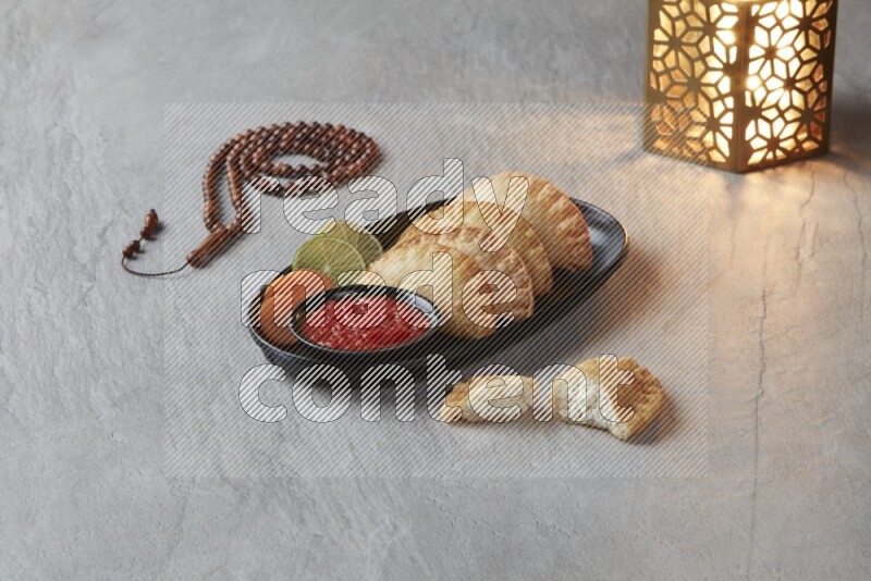 Four fried sambosas in an oval shaped black plate, beside a cut cheese sambosa, a brown misbaha and a golden lantern on a gray background