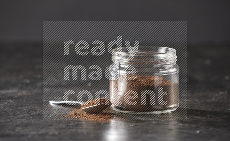 A glass jar full of cloves powder with a metal spoon on a textured black flooring