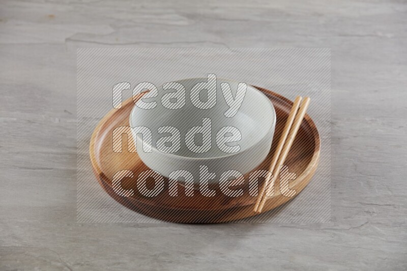 white pottery round bowl on top of brown wood round plate and wood chopsticks, on grey textured countertop