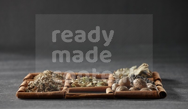 9 squares of cinnamon sticks full of cardamom in the middle surrounded by nutmeg, black pepper, bay laurel leaves, allspice, cumin, white pepper, dried basil and salt on black flooring