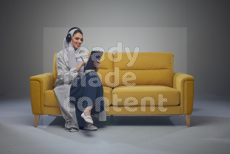 A Saudi woman wearing a light gray Abaya and head scarf sitting on a yellow sofa and using her phone while wearing headphones eye level on a grey background