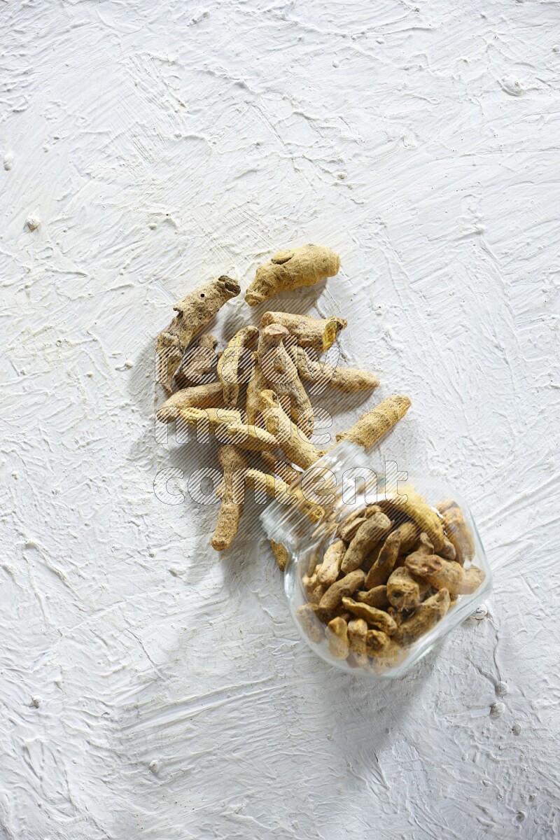 A flipped glass spice jar full of dried turmeric fingers and fell out it on textured white flooring