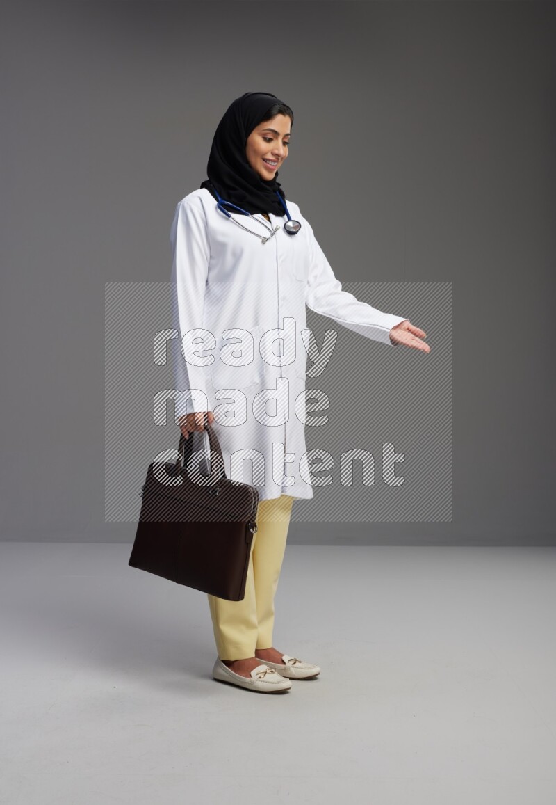 Saudi woman wearing lab coat with stethoscope standing holding bag on Gray background
