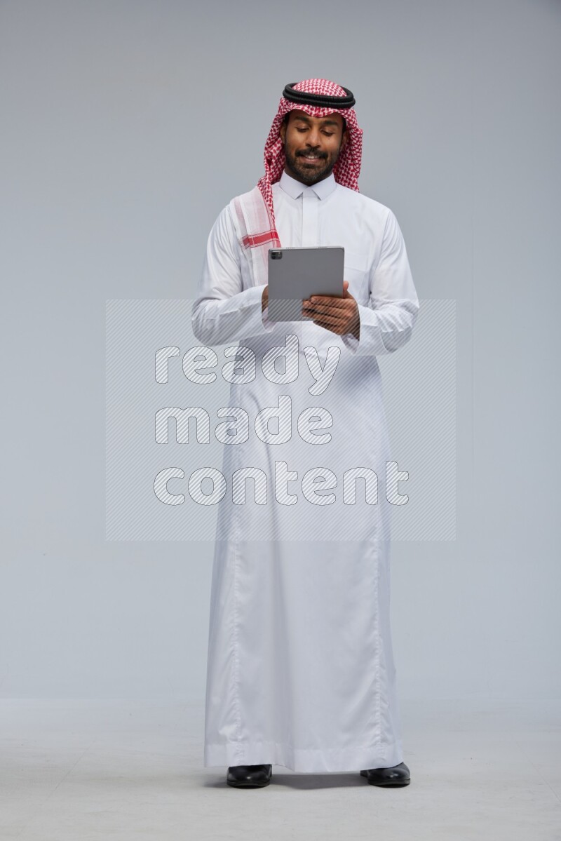 Saudi man Wearing Thob and shomag standing working on tablet on Gray background