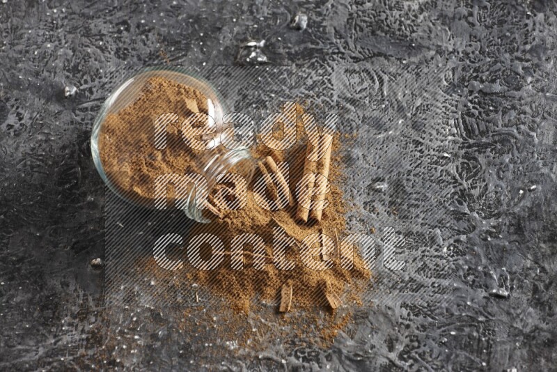 Flipped glass herbs jar full of cinnamon powder with cracked cinnamon sticks on a textured black background