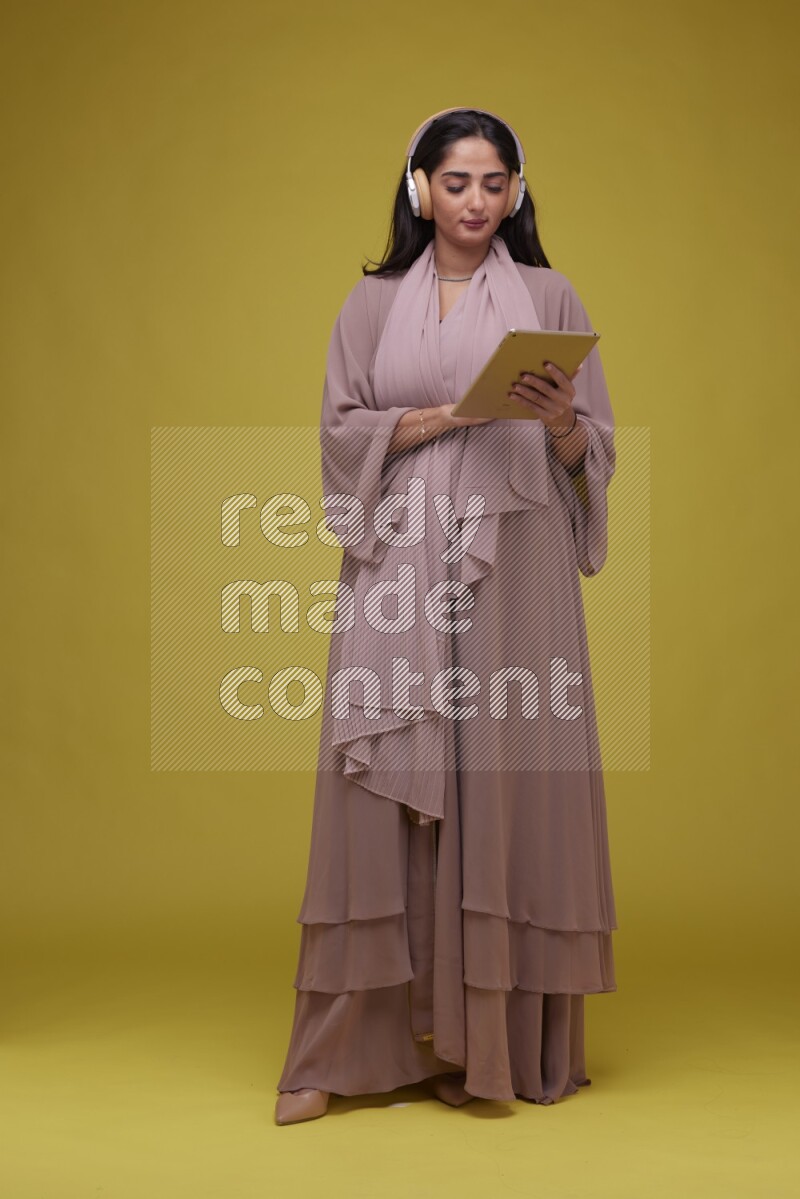 A woman Holding an Ipad with Headphone on a Yellow Background wearing Brown Abaya
