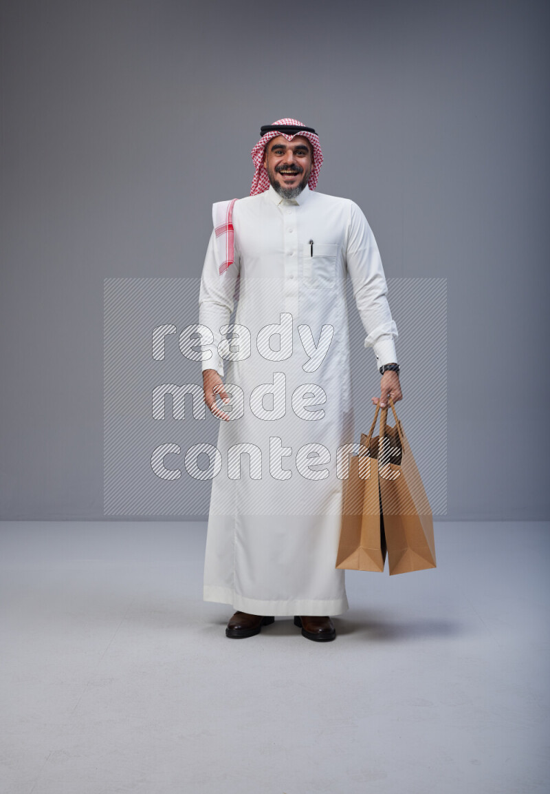 Saudi man Wearing Thob and red Shomag standing holding shopping bag on Gray background