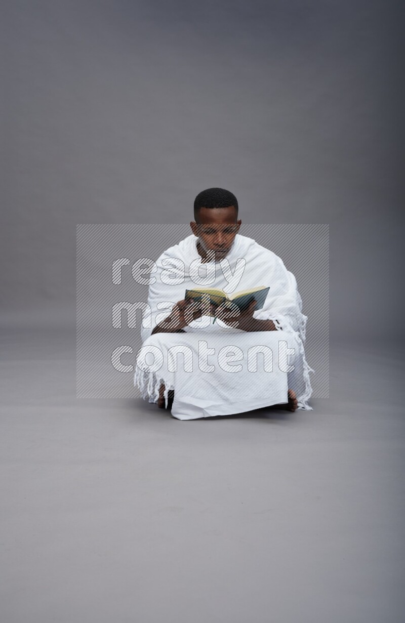 A man wearing Ehram sitting on floor reading quran on gray background