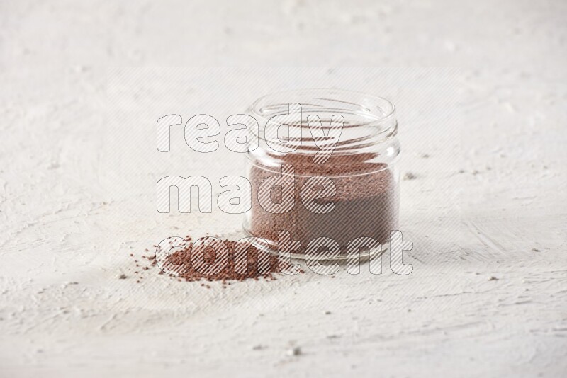 A glass jar full of garden cress seeds with more seeds spread on a textured white flooring