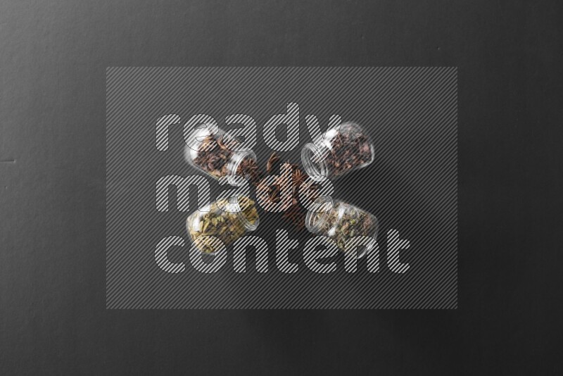 4 glass spice jars full of cloves, cardamom, basil and star anise on black flooring
