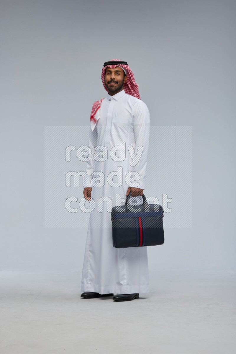 Saudi man Wearing Thob and shomag standing holding bag on Gray background