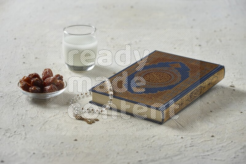 Quran with dates, prayer beads and different drinks all placed on textured white background