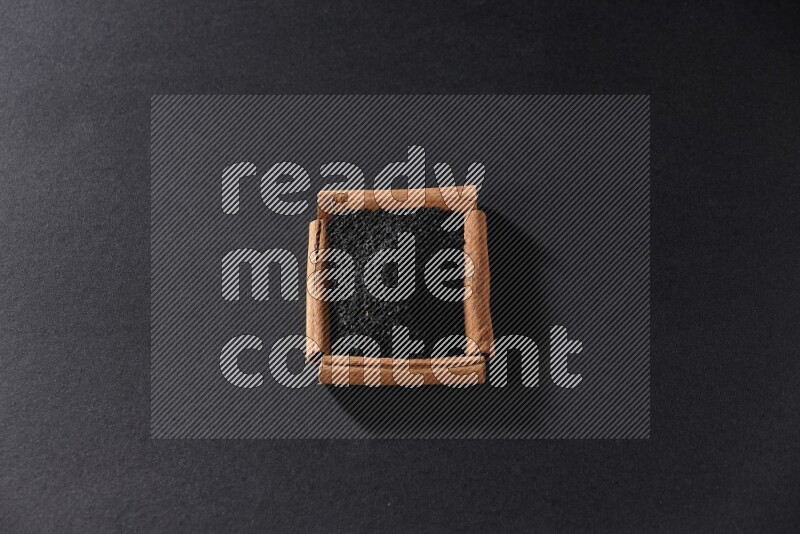 أربعة من أعواد القرفة مكونة شكل مربع بداخلها حبة البركة علي خلفية سوداء