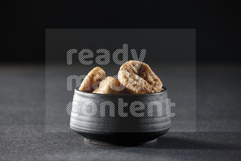 A black pottery bowl full of dried figs on a black background in different angles