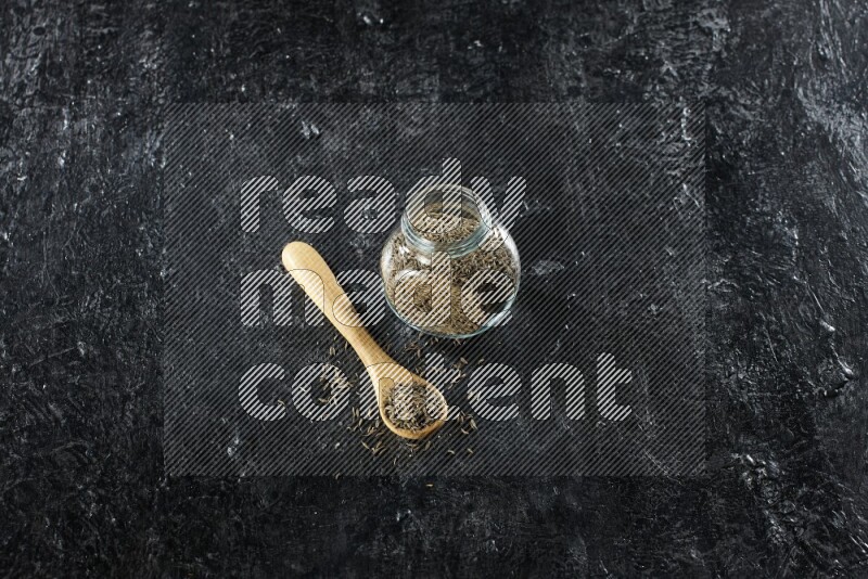 A glass spice jar and a wooden spoon full of cumin seeds on a textured black flooring
