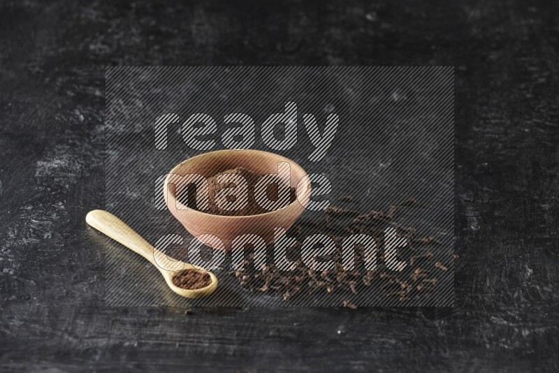 A wooden bowl and wooden spoon full of cloves powder with spreaded cloves on textured black flooring
