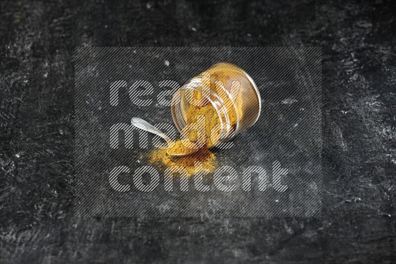 A flipped glass jar and a metal spoon full of turmeric powder and powder spilled out of it on textured black flooring