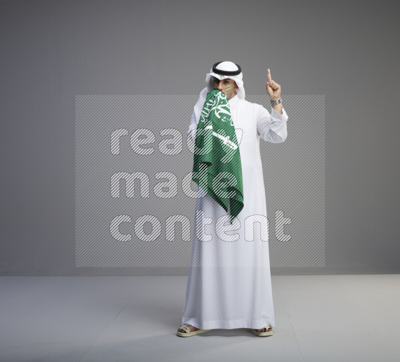 A Saudi man standing wearing thob and white shomag with face painting kissing big Saudi flag on gray background