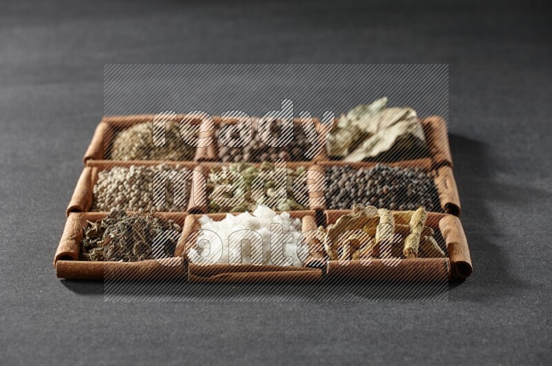 9 squares of cinnamon sticks full of cardamom in the middle surrounded by turmeric, black peppers, bay laurel, allspice, cumin, white peppers, basil and salt on black flooring