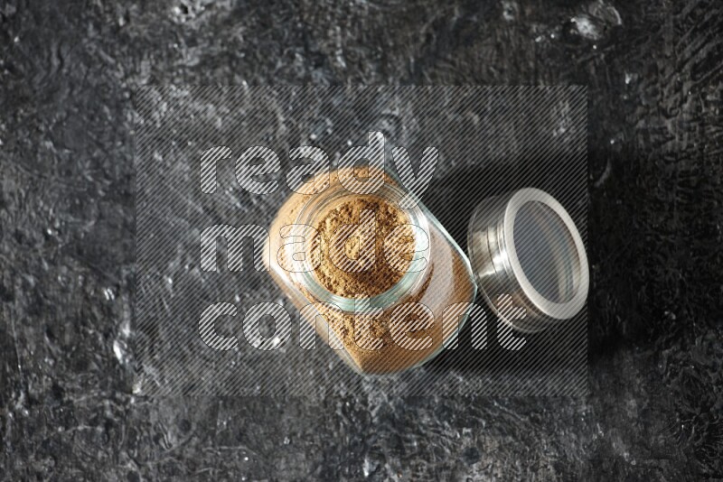 A glass spice jar full of allspice powder on a textured black flooring