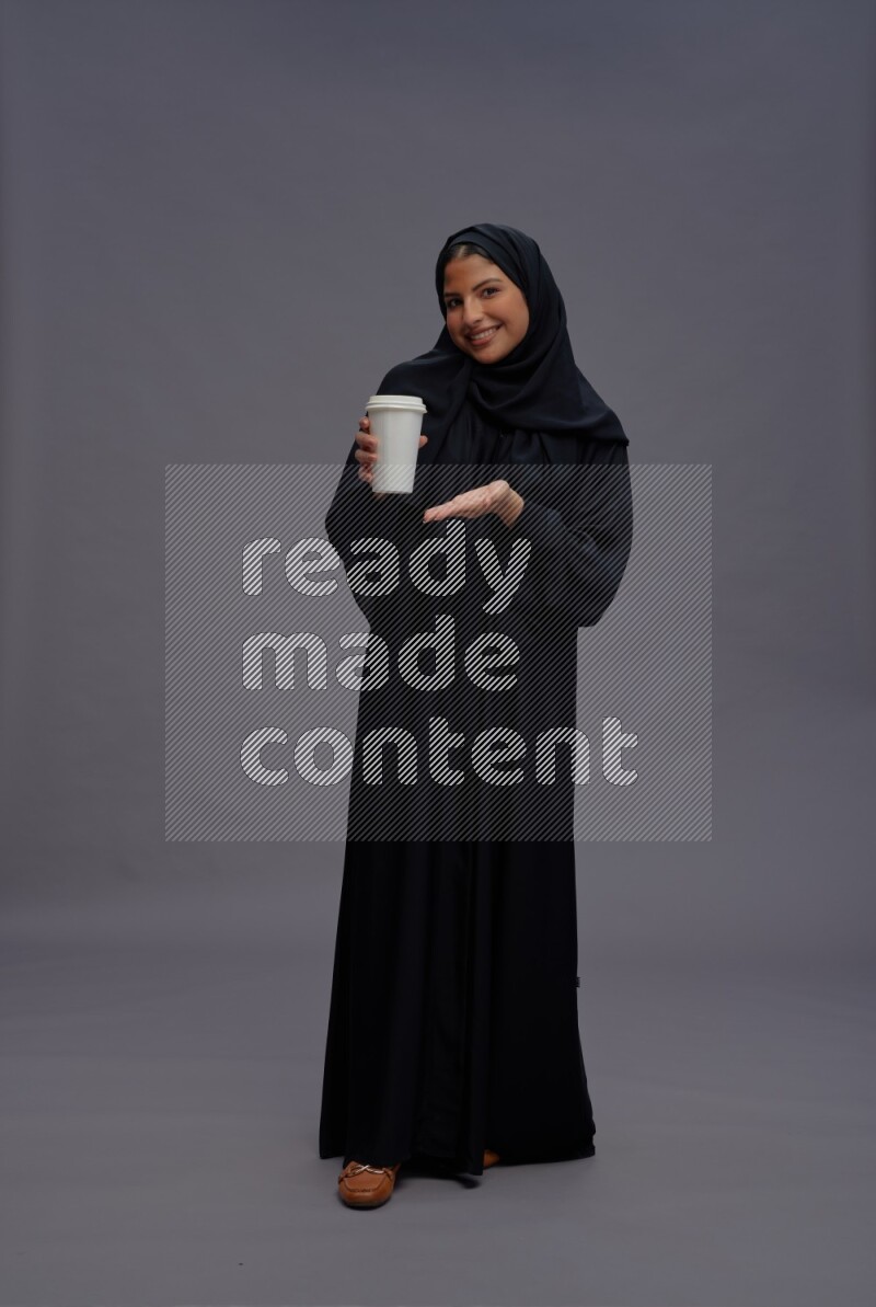 Saudi woman wearing Abaya standing holding paper cup on gray background