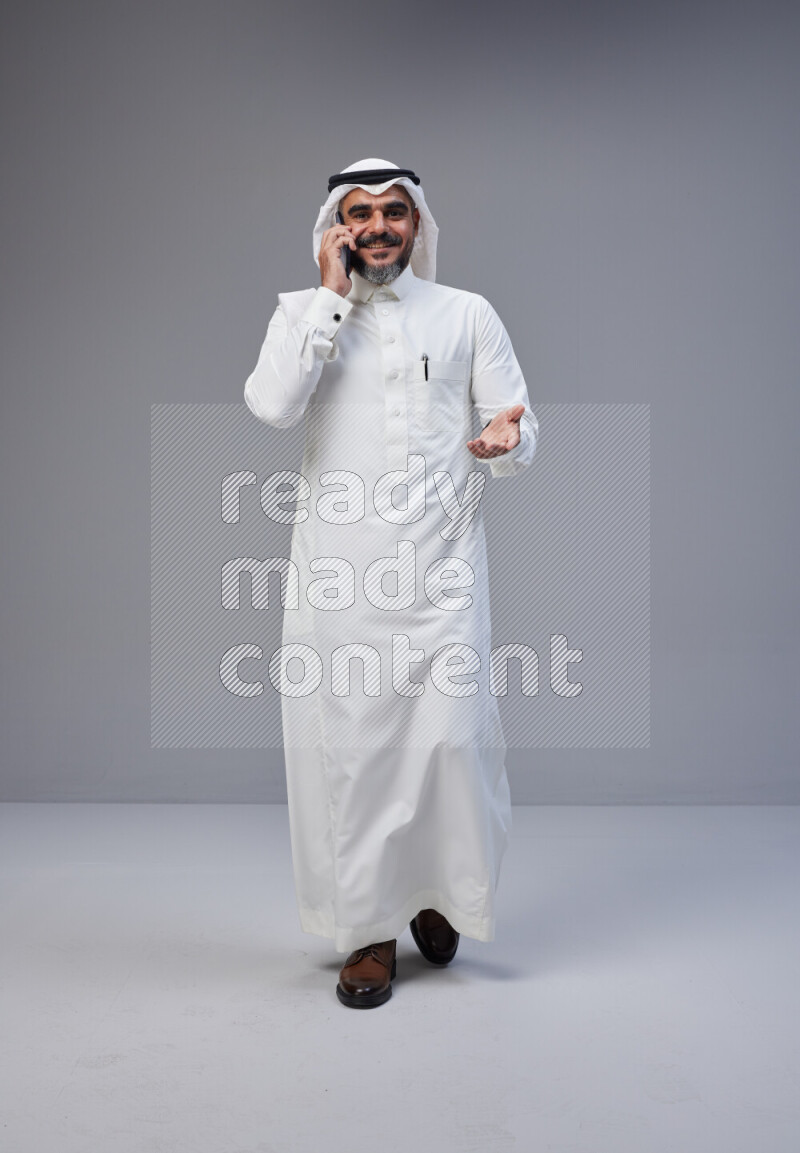 Saudi man Wearing Thob and white Shomag standing talking on phone on Gray background