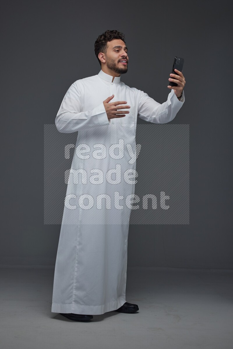 A Saudi man wearing Thobe and taking selfie with his phone making different poses eye level on a gray background