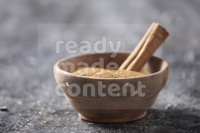 Wooden bowl full of cinnamon powder and a cinnamon stick on a textured black background