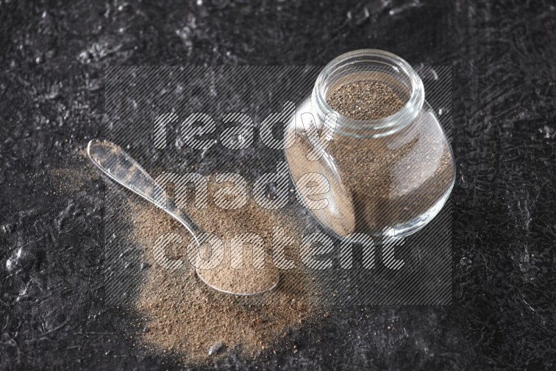 A glass spice jar full of black pepper powder and a metal spoon full of powder on textured black flooring
