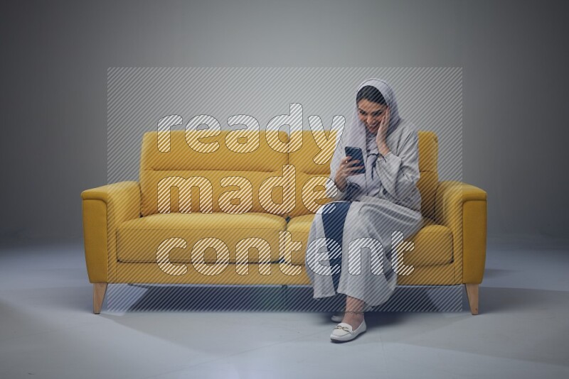A Saudi woman wearing a light gray Abaya and white head scarf sitting on a yellow sofa and using her phone eye level on a grey background