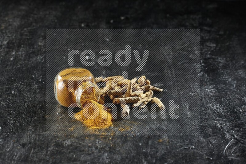 A flipped glass spice jar full of turmeric powder and powder spilled out of it with dried whole fingers on textured black flooring