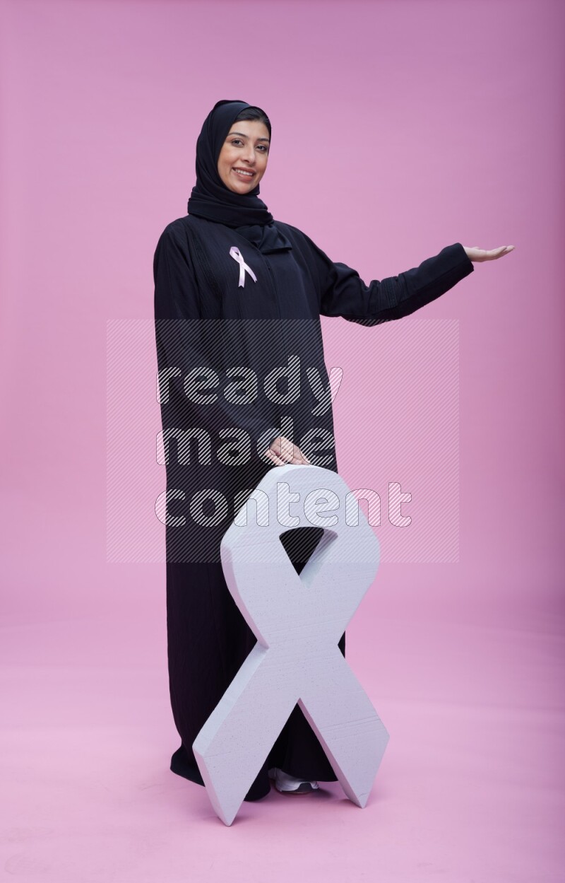 Saudi woman wearing pink ribbon on abaya standing holding awareness ribbon on plain pink background