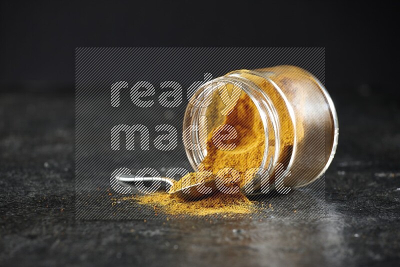 A flipped glass jar and a metal spoon full of turmeric powder and powder spilled out of it on textured black flooring