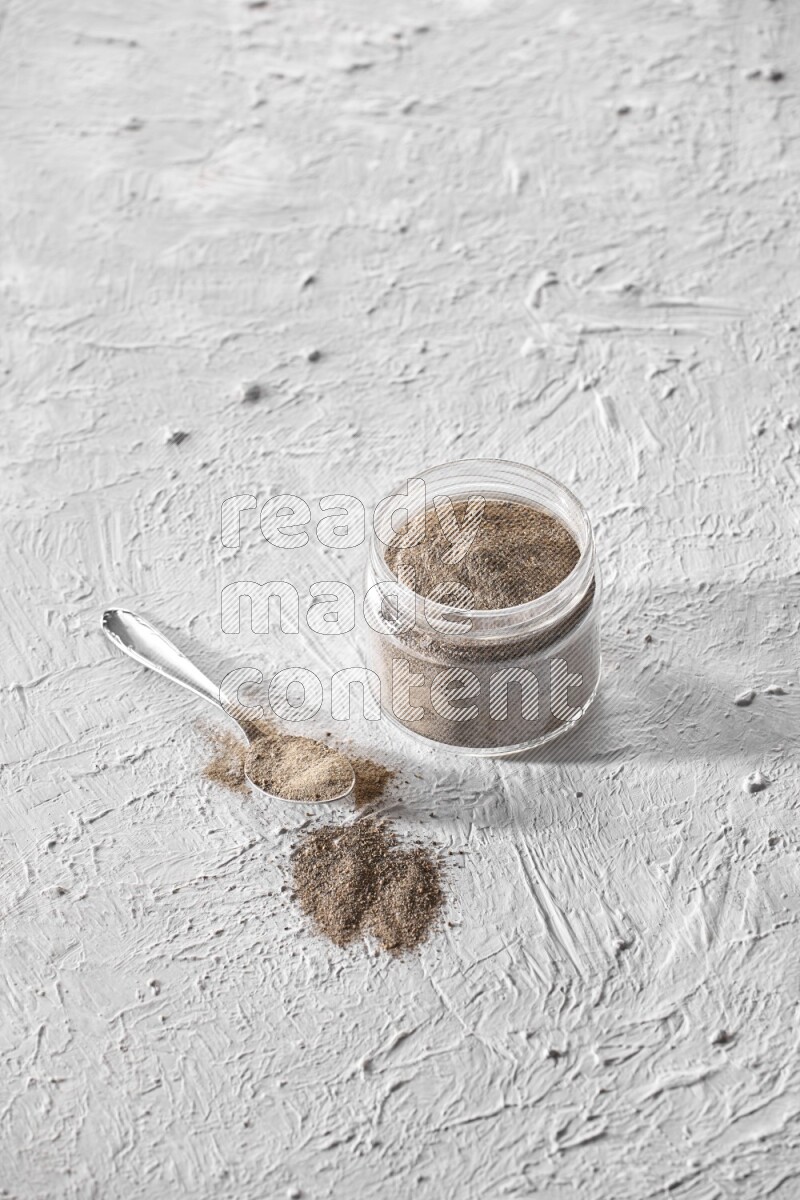 A glass jar full of black pepper powder and a metal spoon full of powder on a textured white flooring