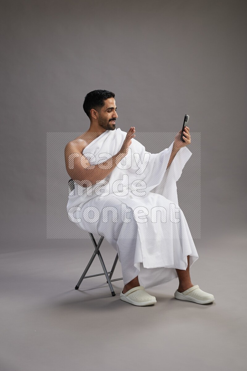 A man wearing Ehram sitting on chair taking selfie on gray background
