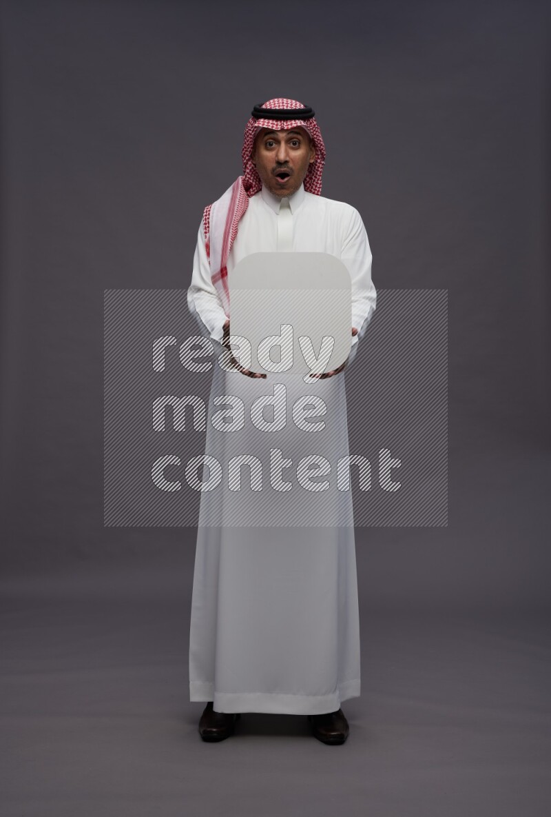 Saudi man wearing thob and shomag standing holding social media sign on gray background
