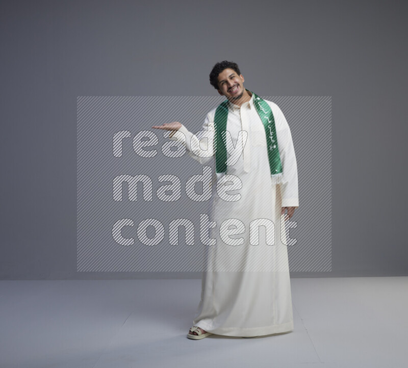 A Saudi man standing wearing thob and Saudi flag scarf on gray background