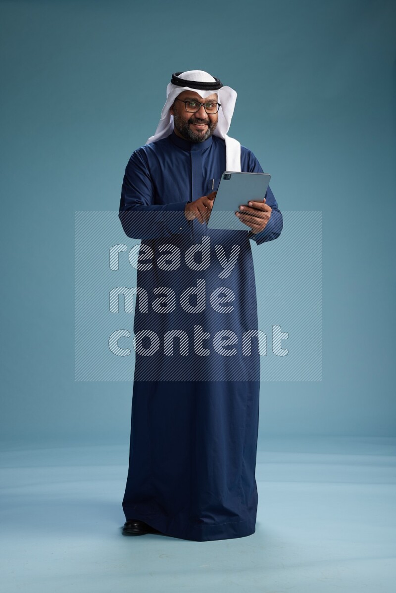 Saudi Man with shimag Standing holding ATM card while working on tablet on blue background