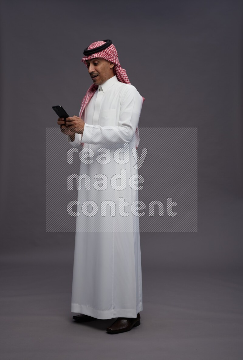 Saudi man wearing thob and shomag standing texting on phone on gray background