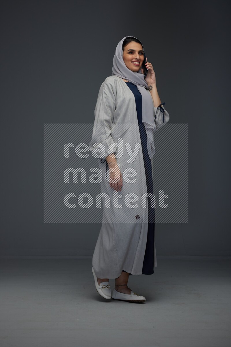 A Saudi woman wearing a light gray Abaya and head scarf standing and talking in the phone eye level on a grey background