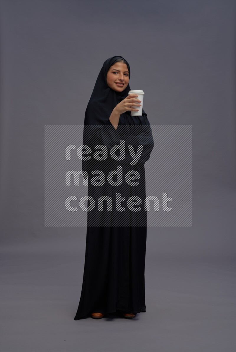 Saudi woman wearing Abaya standing holding paper cup on gray background