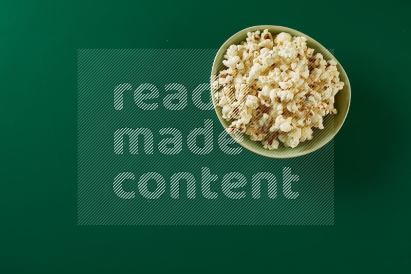 A green ceramic bowl full of popcorn on a green background in a top view shot