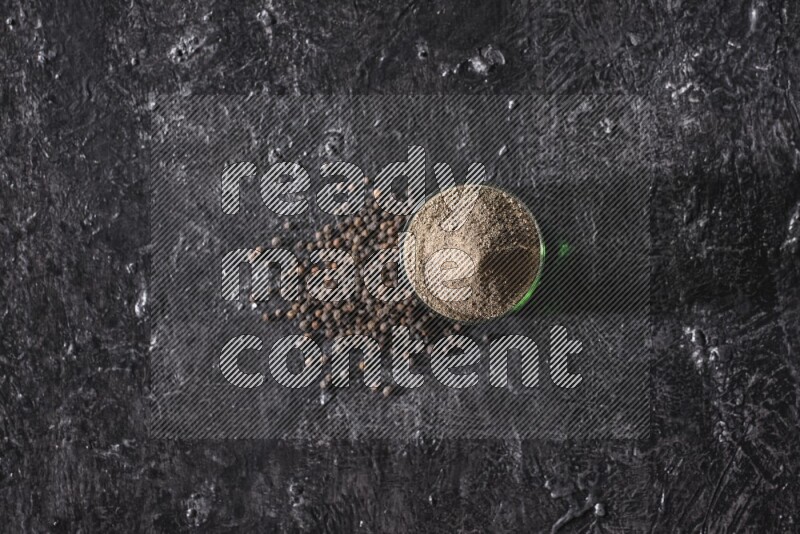 A glass cup full of black pepper powder with beads beside it on a textured black flooring