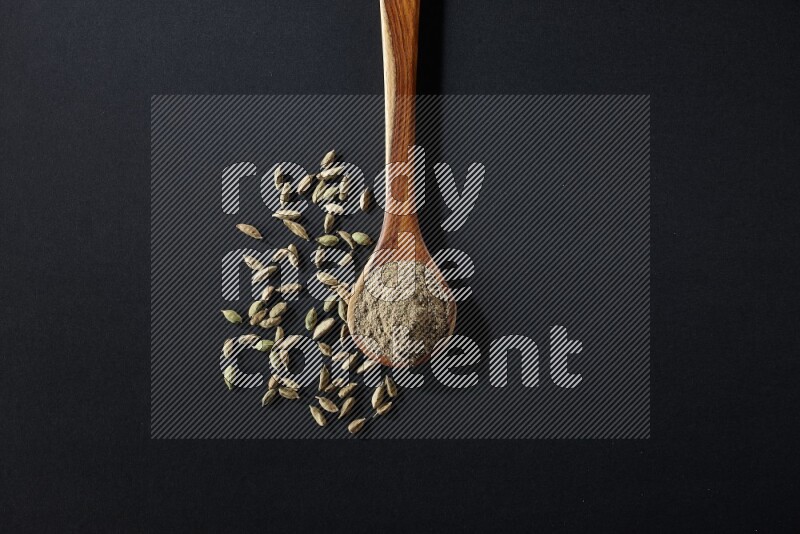 A wooden ladle full of cardamom powder and cardamom seeds beside it on black flooring