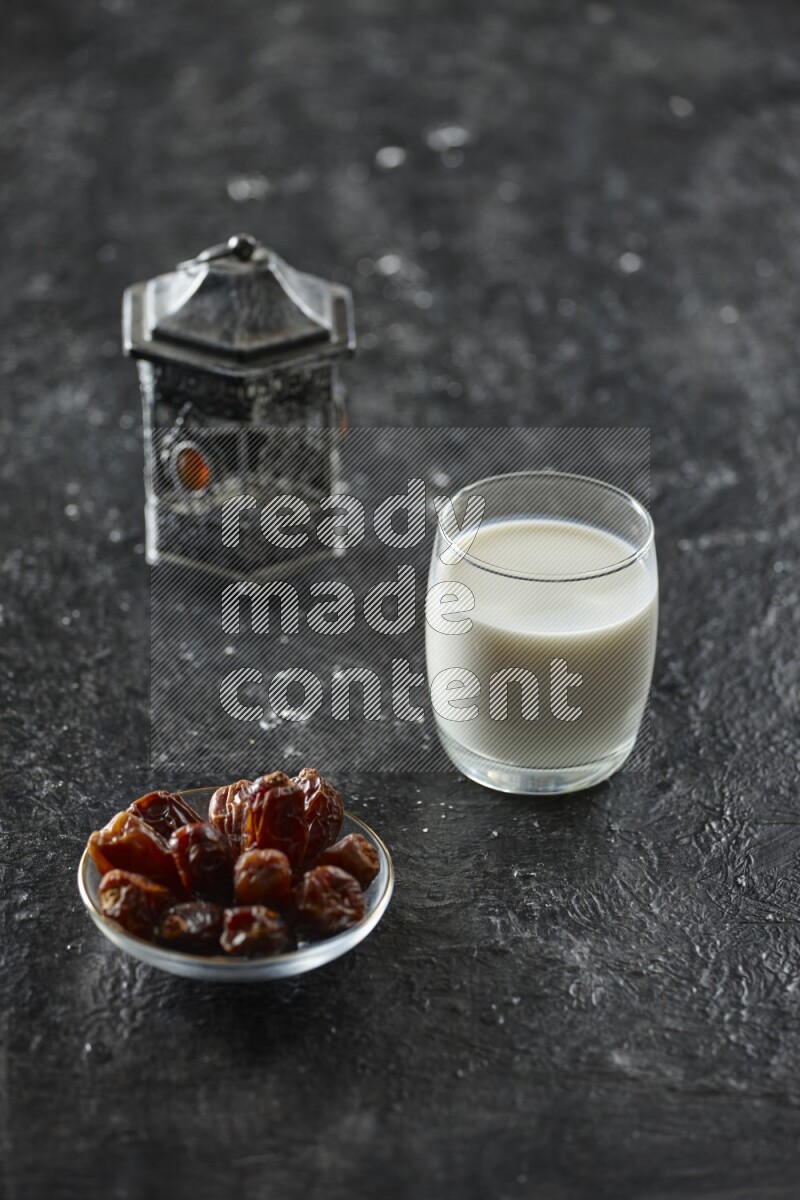 A silver lantern with different drinks, dates, nuts, prayer beads and quran on textured black background