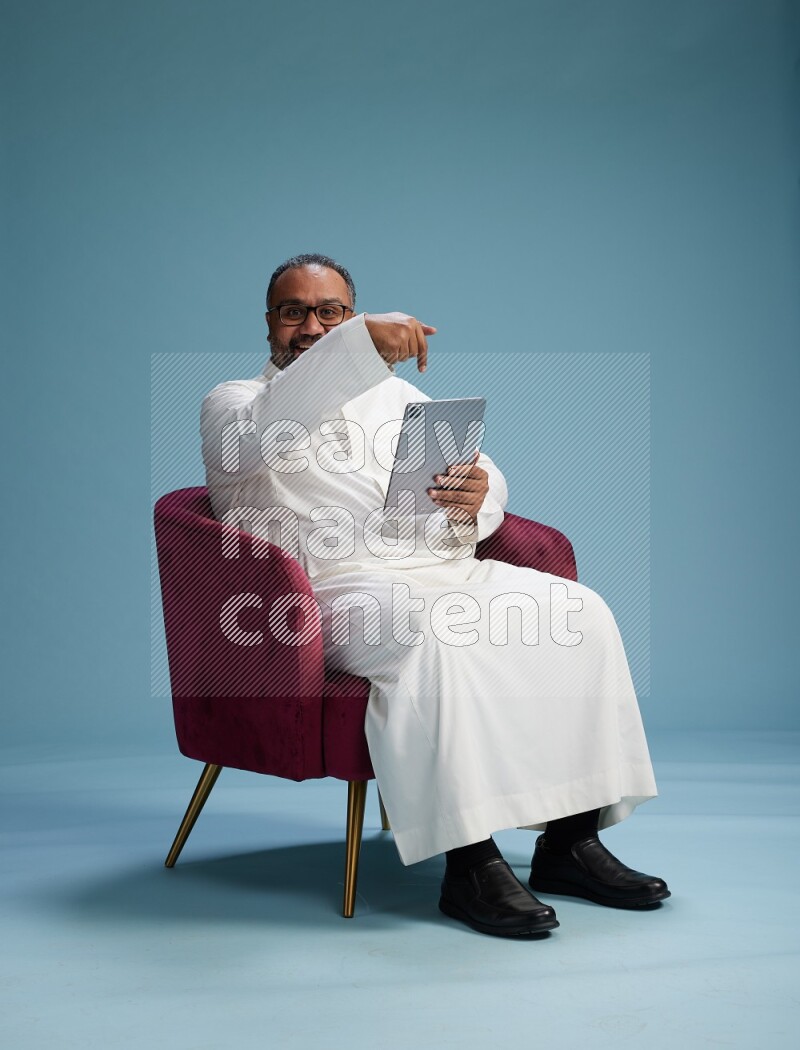Saudi Man without shimag sitting on chair working on tablet on blue background