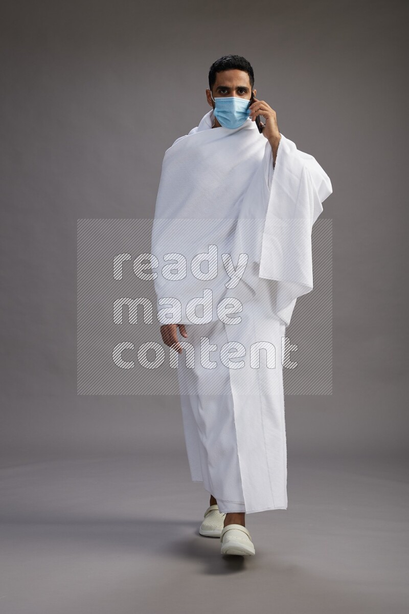 A man wearing Ehram with face mask Standing talking on phone on gray background