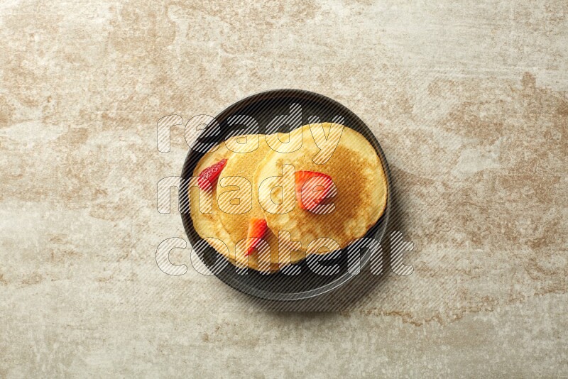 Three stacked strawberry pancakes in a black plate on beige background